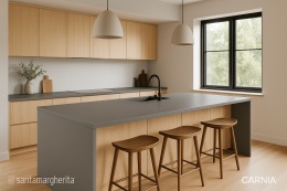 Minimalistyczna kuchnia z wyspą i blatami z konglomeratu Santamargherita Carnia, jasne drewniane szafki i hokery.