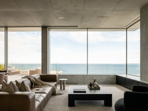 Minimalistyczny salon z panoramicznym widokiem na morze i betonowymi wykończeniami ścian.