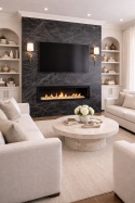 Salon z kominkiem w ścianie ze spieku Nero Greco, kremowe miękkie sofy i elegancki stolik kawowy w jasnym kamieniu.