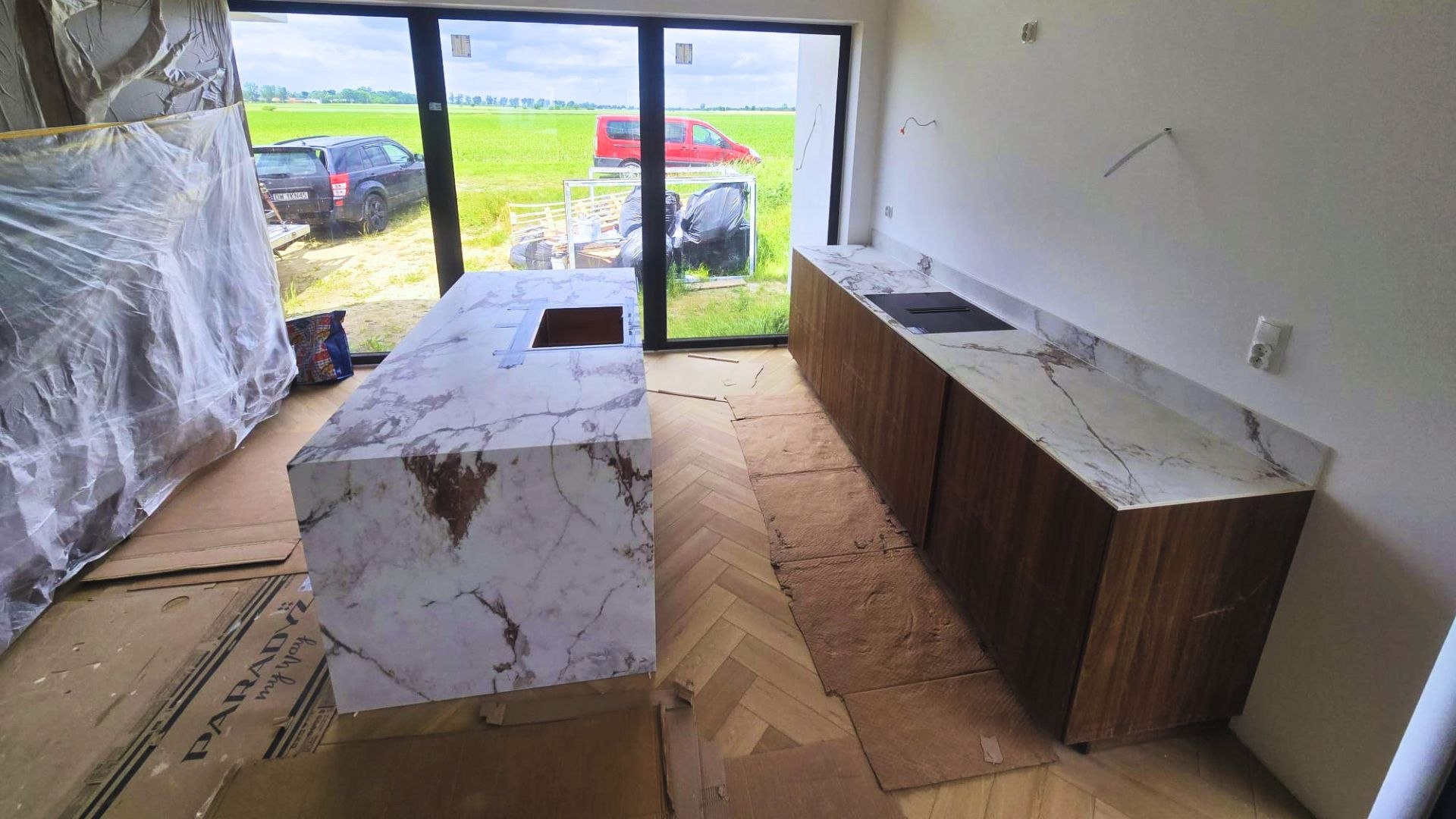 Widok kuchni z dużą wyspą i zabudową przyścienną w spieku Marble Capraia. Blaty zestawione z drewnianymi frontami i naturalnym światłem z panoramicznego okna.
