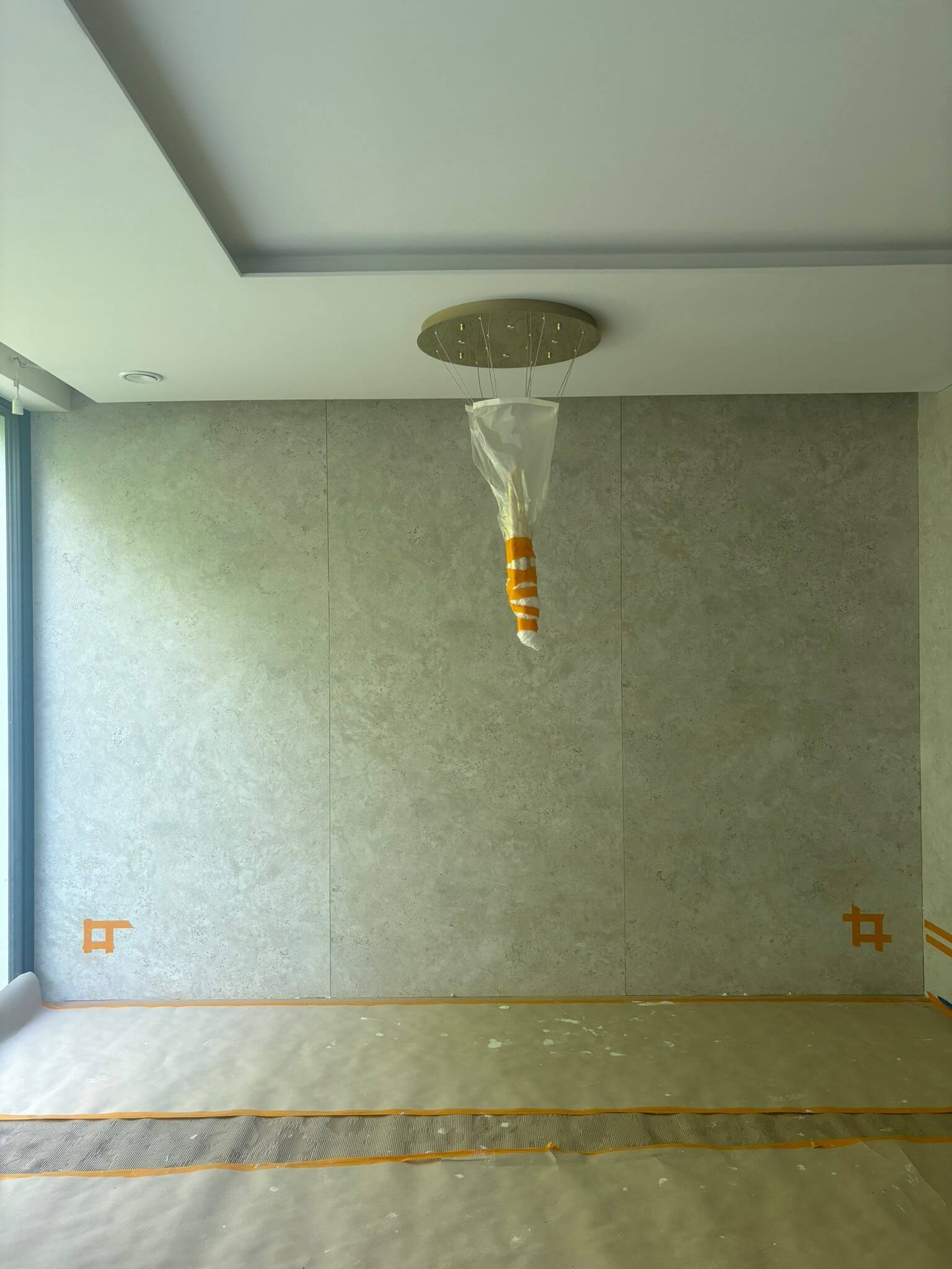 Etap prac wykończeniowych w nowoczesnym wnętrzu – ściana z zamontowanymi płytami Dekton Sabbia, sufit z podwieszeniem, złoty żyrandol w folii ochronnej, zabezpieczona podłoga