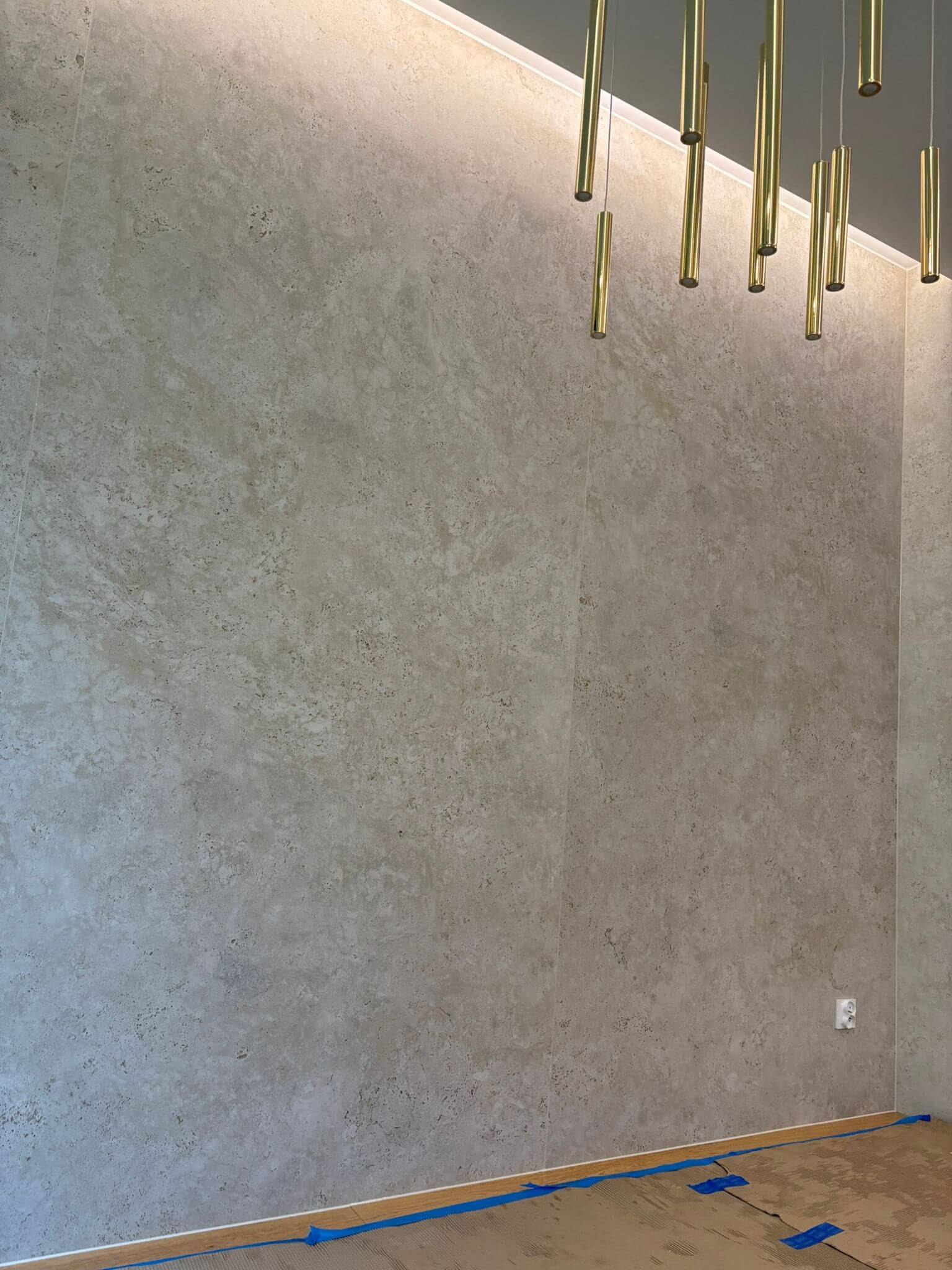 Nowoczesna ściana z płyt Dekton Sabbia w trakcie realizacji – widoczna struktura spieku, oświetlenie liniowe przy suficie i złote lampy wiszące, zabezpieczona podłoga
