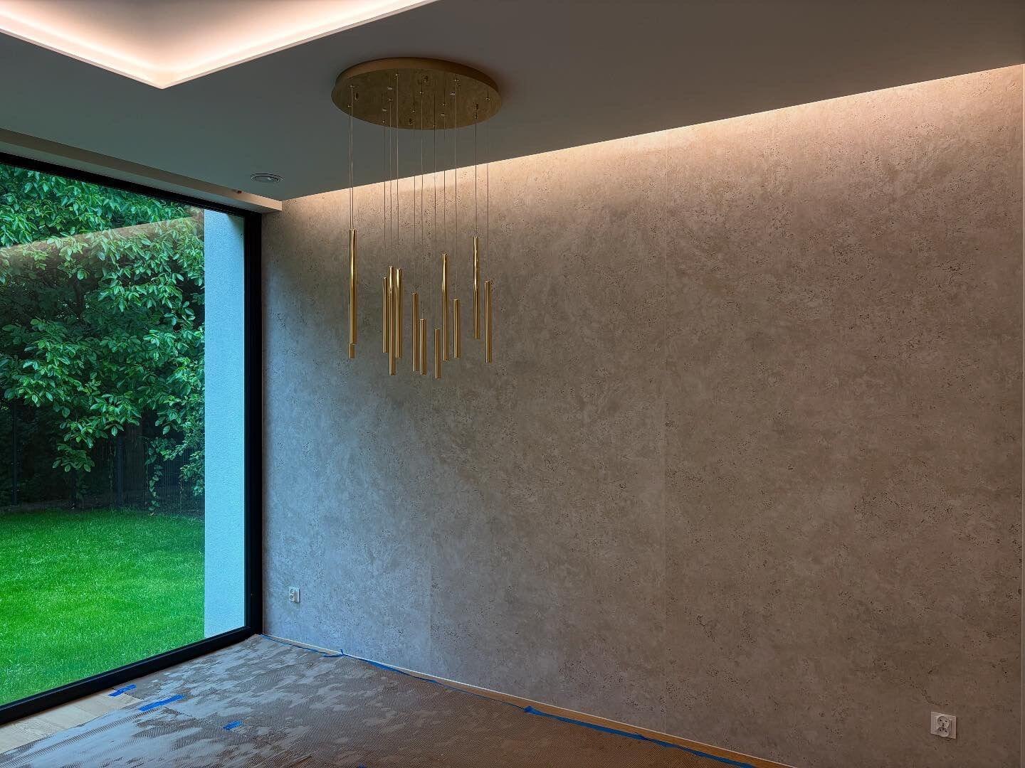 Nowoczesne wnętrze z dużym oknem i ścianą wykończoną spiekiem kwarcowym Dekton Sabbia; złoty żyrandol i oświetlenie liniowe podkreślają naturalną strukturę materiału